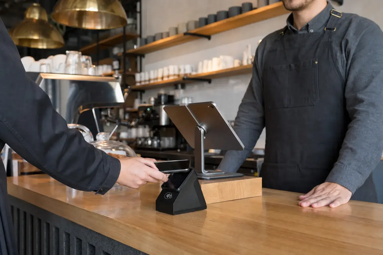 A customer making a contactless payment by tapping a smartphone on a matte-black card terminal at a modern cafe counter, with a barista and a point-of-sale tablet